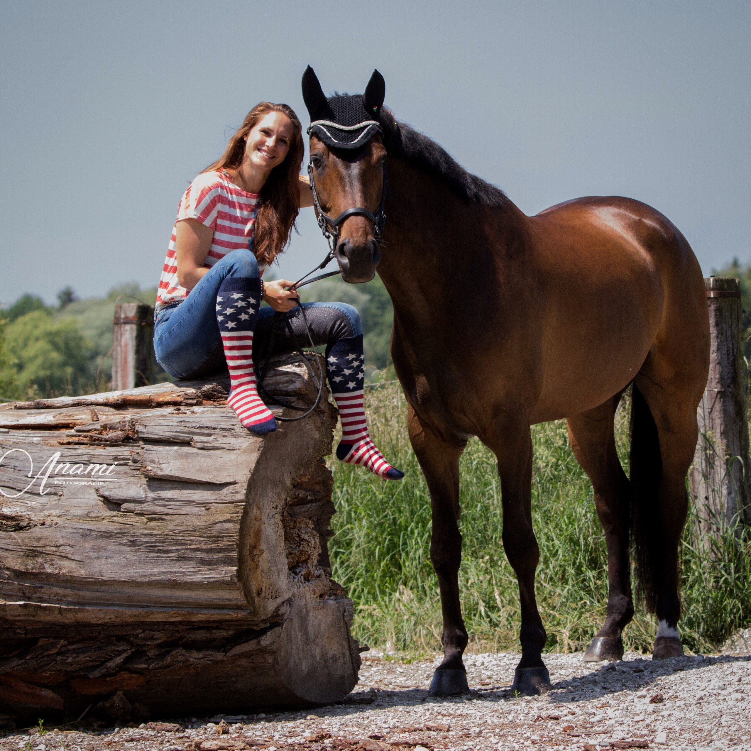 Pferd neben Baumstamm, auf dem die Besitzerin in USA-Ringelsocken sitzt und lacht